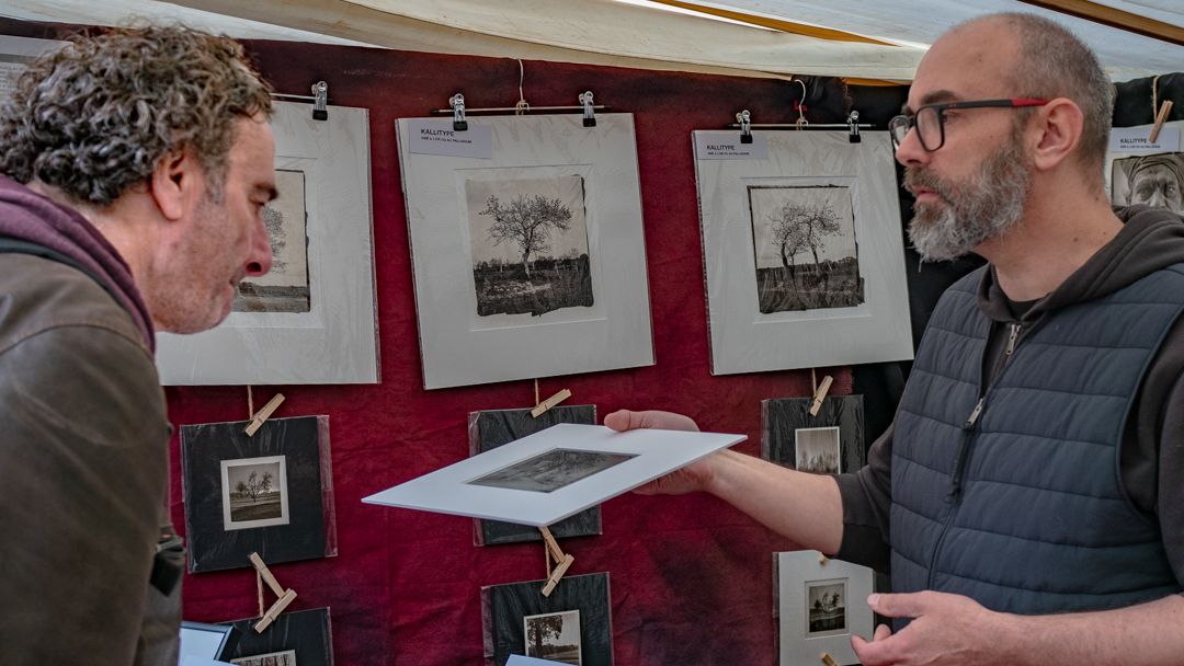 A stallholder at the artists' market holds up a print for a visitor to inspect, with more prints clipped to a red backdrop behind them.