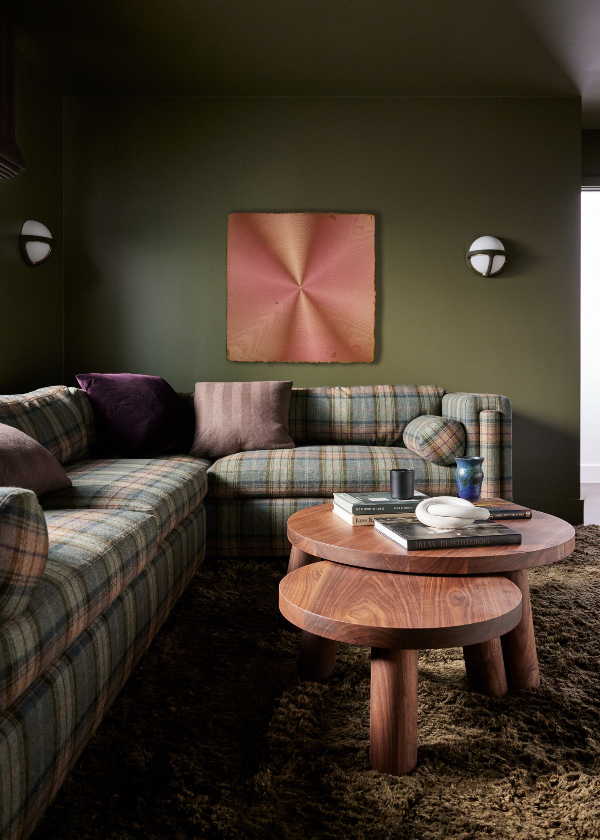 A cozy living room with dark green walls, a plaid sofa, two wooden round coffee tables, a shaggy rug, and pink artwork on the wall.