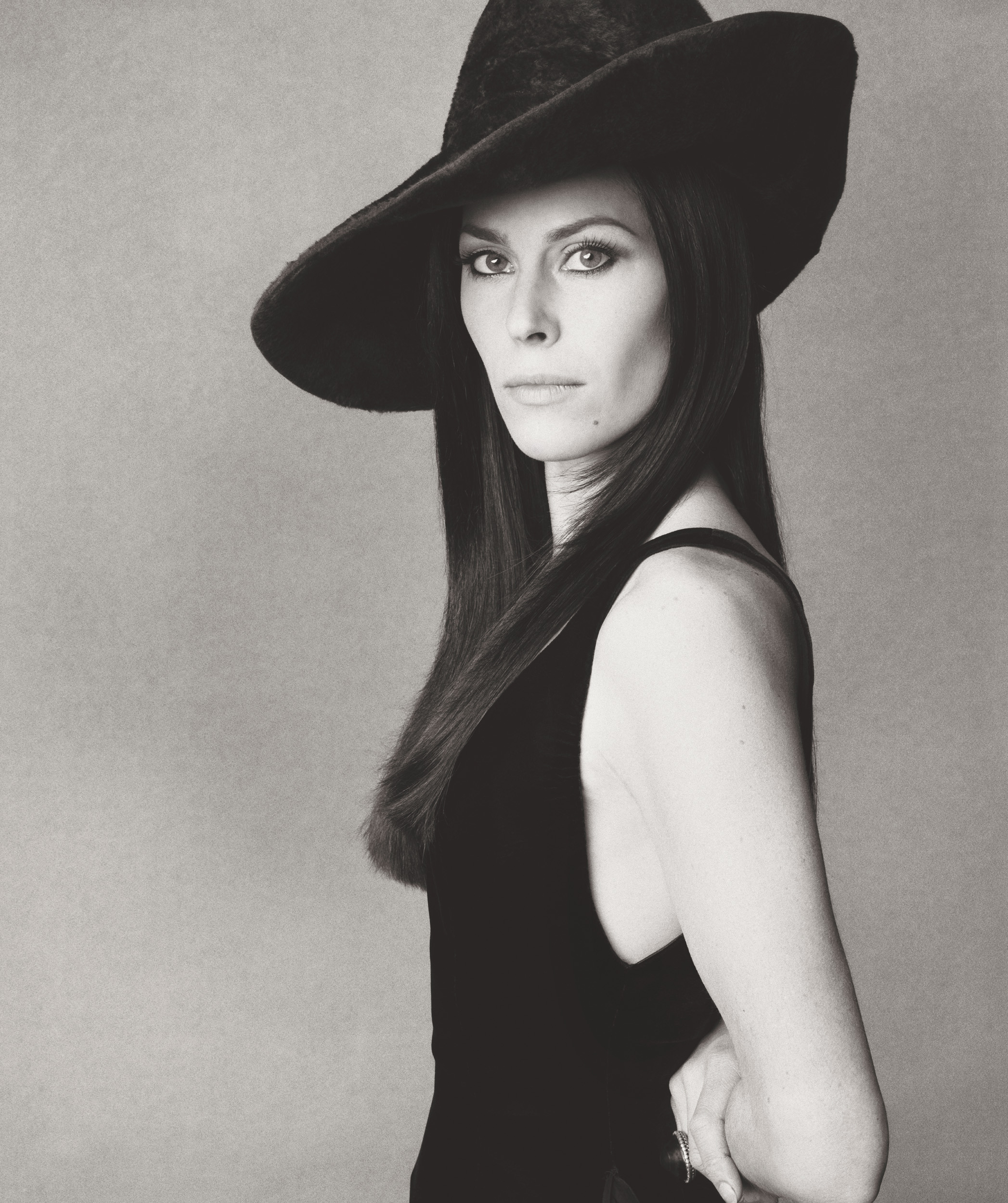 A black and white profile portrait of a woman with long, dark hair wearing a wide-brimmed black hat and a sleeveless black dress.