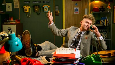 A man in a suit sits with his legs up on a desk, holding a telephone to his ear in a messy office