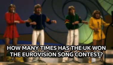 Bucks Fizz perform on stage at Eurovision, dancing behind mics on stands. The girls wear long sleeved tops and skirts, one red and one yellow. The boys in the centre wear white pants and navy jumpers with red and yellow stripes on the arms