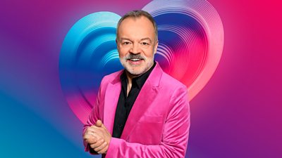 A man in a black shirt and hot pink blazer smiles to camera with his hands clasped. He's set in front of a pulsing blue, pink and purple heart.