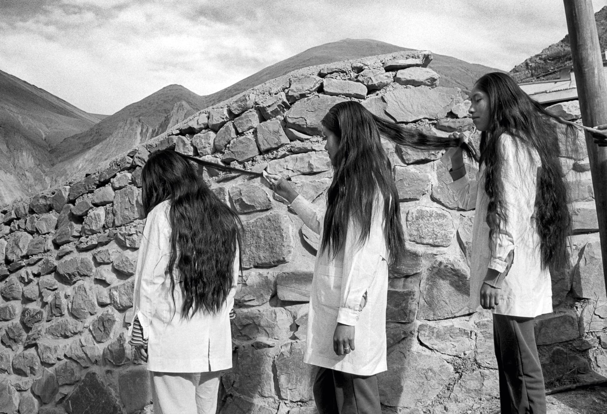 A black-and-white photo of three young women with long black hair, near a stone wall