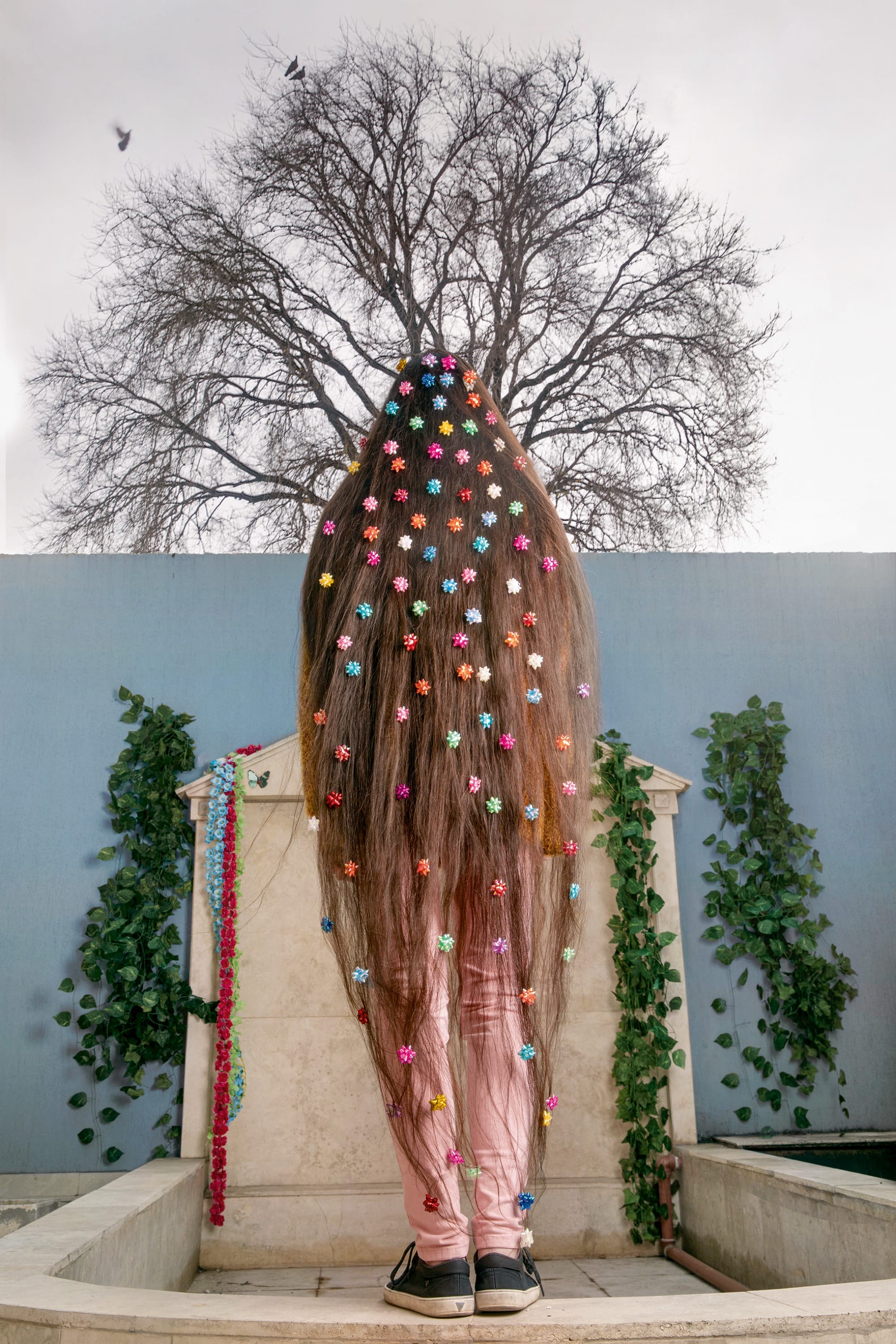 A young woman with very long hair stands facing away from the camera, with colorful dots in her hair