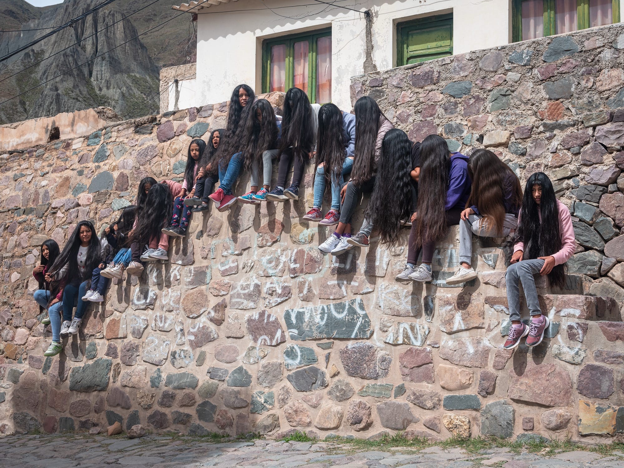 A group of young women with very long hair sit along a stone wall in a line