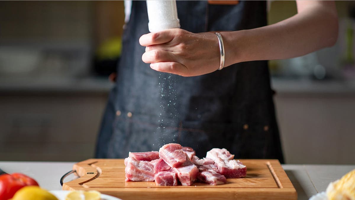 A woman in an apron salts a raw piece of meat on a cutting board.