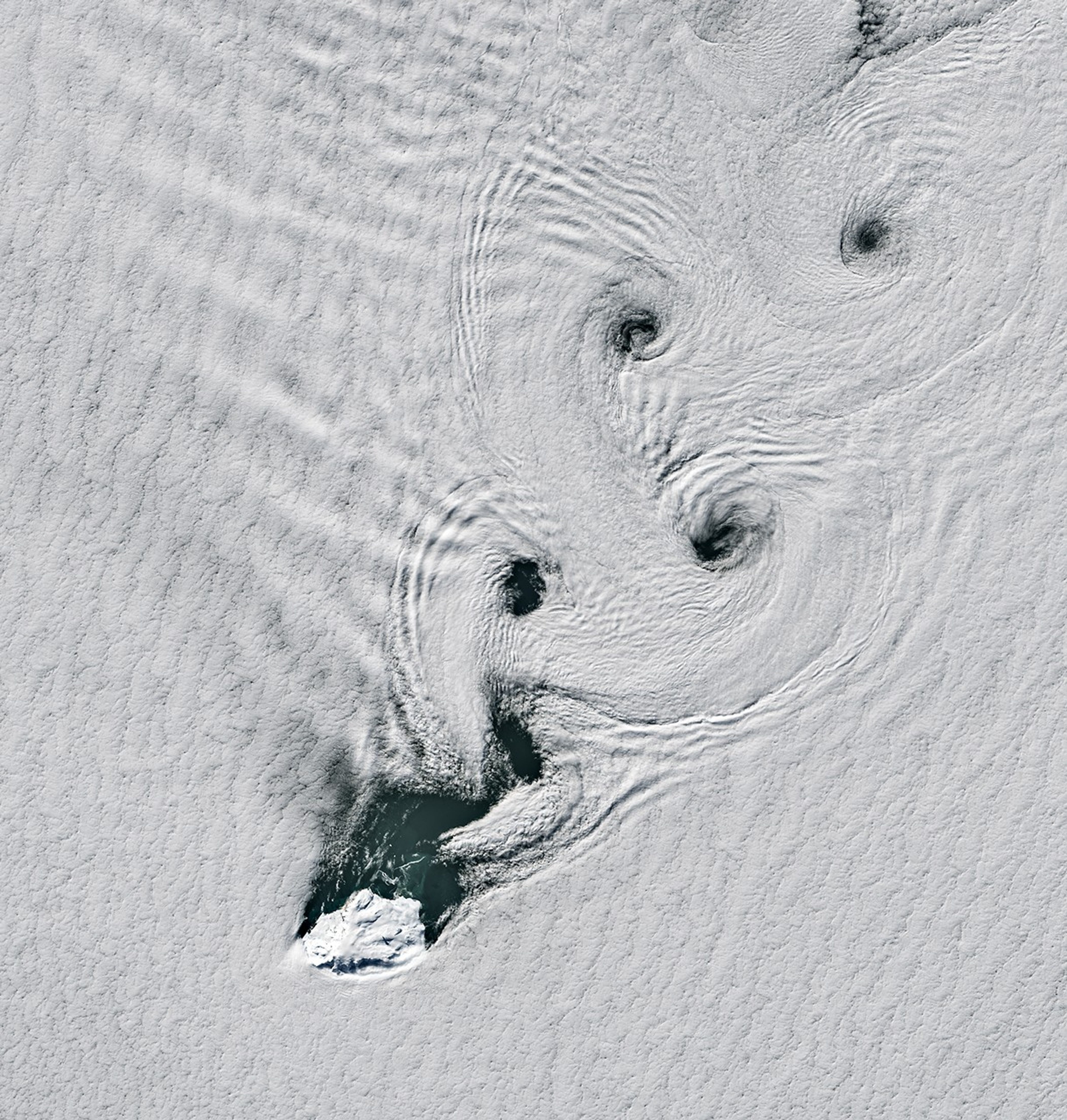 Parallel lines of cloud vortices appear downwind of a small, ice-covered island.