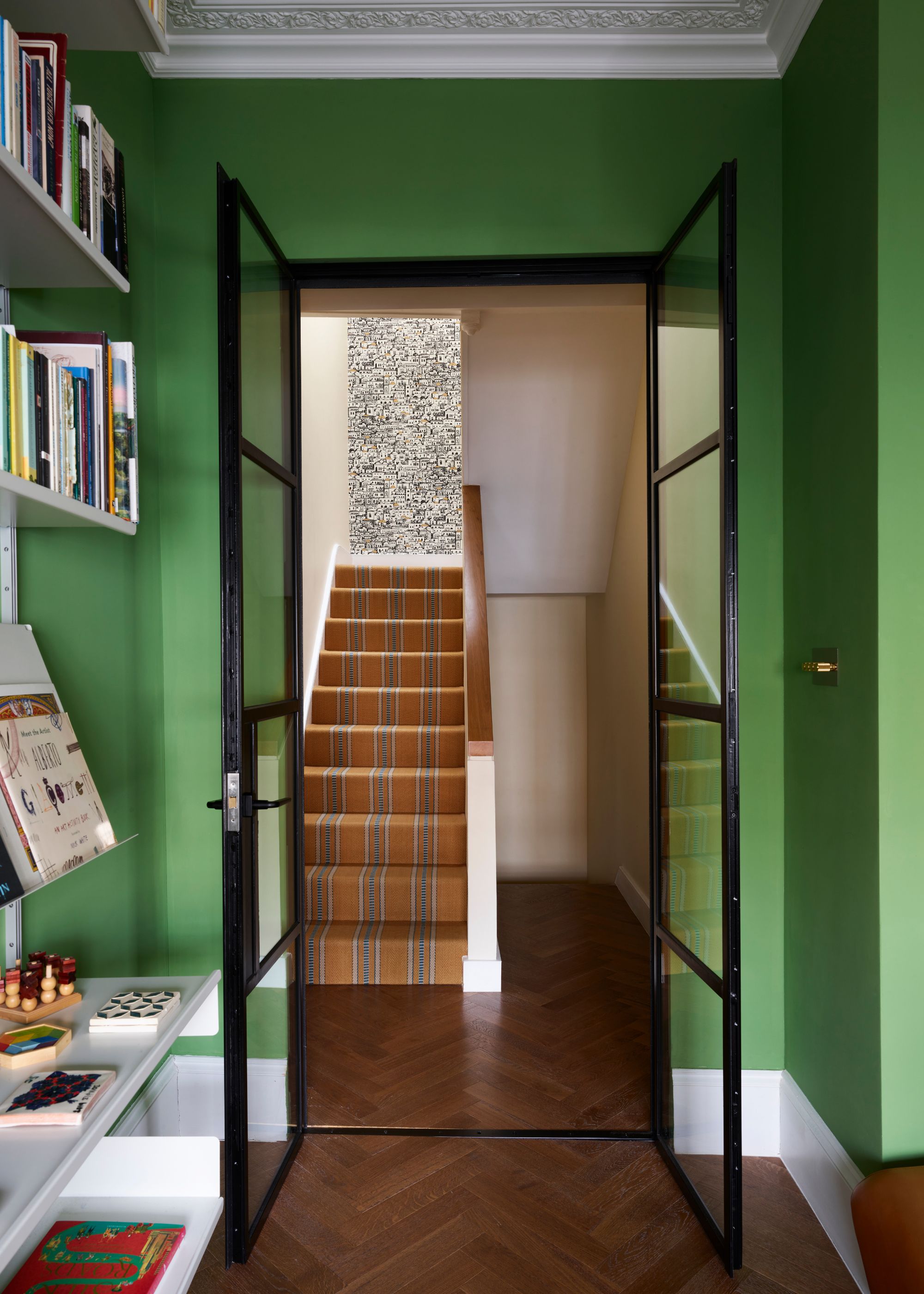 A living room with bright green walls, white woodwork, white shelving units, and parquet flooring. Black-framed glass doors leading into a hallway with beige walls and a mustard staircase runner.
