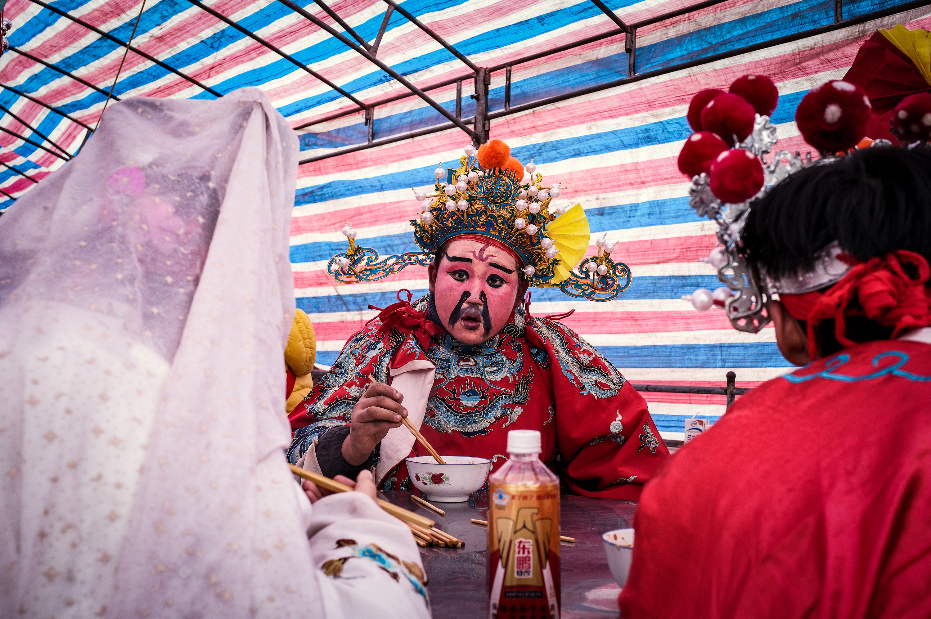 A performer in elaborate red costume with gold-embroidered headdress and bright face paint eats from a small bowl beneath a colorful striped tent.