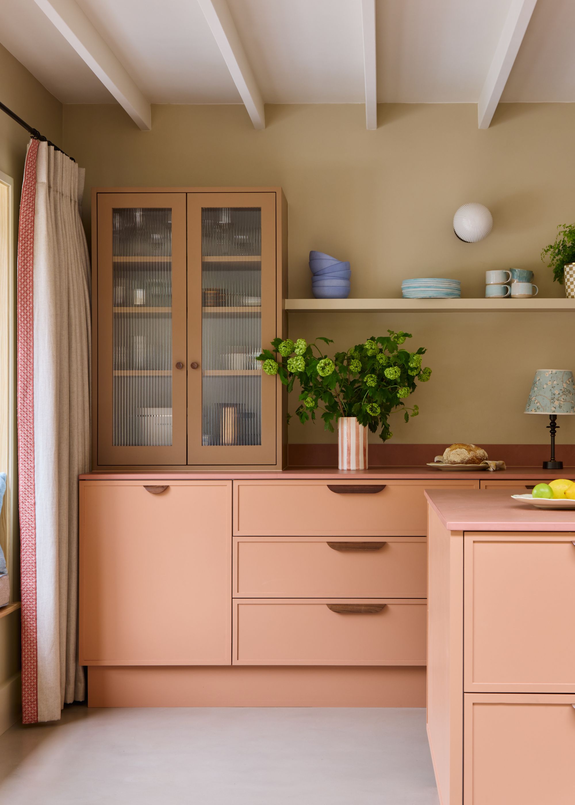 A modern kitchen with salmon pink cabinets, sandy beige walls, a caramel brown wall cabinet, a pink kitchen island, light grey flooring, and a white ceiling.