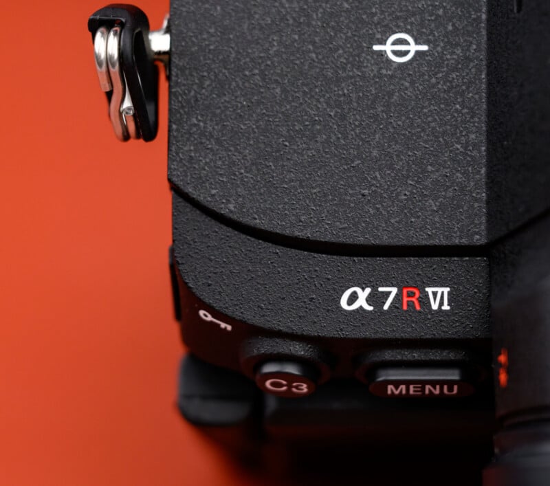 Close-up view of the corner of a black Sony Alpha 7R VI camera body, showing the model name, buttons labeled C3 and MENU, against an orange background.