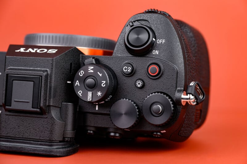 Close-up of the top controls of a Sony digital camera with various dials and buttons visible, set against an orange background.