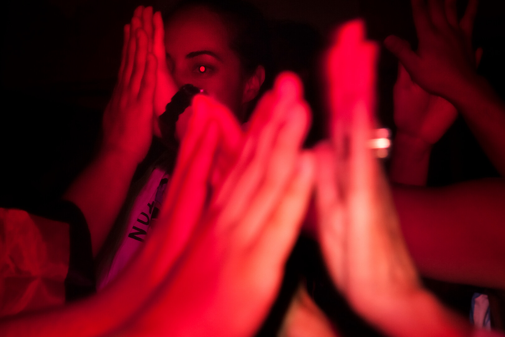 A close-up, red-filtered photograph capturing a woman's intense gaze between several pairs of hands raised in a rhythmic clapping motion.