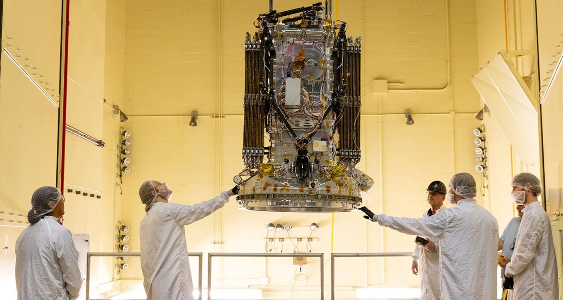 Staff in white lab coats observe a black and gray spacecraft in a room with warm, yellow light.