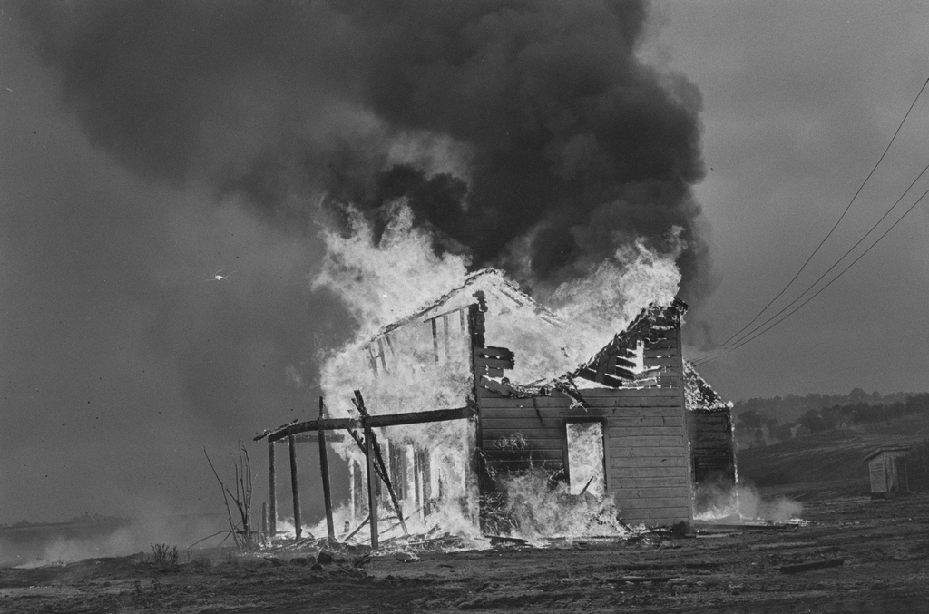 A wooden two-storey building is fully engulfed in flames, sending a thick column of black smoke into a grey sky.