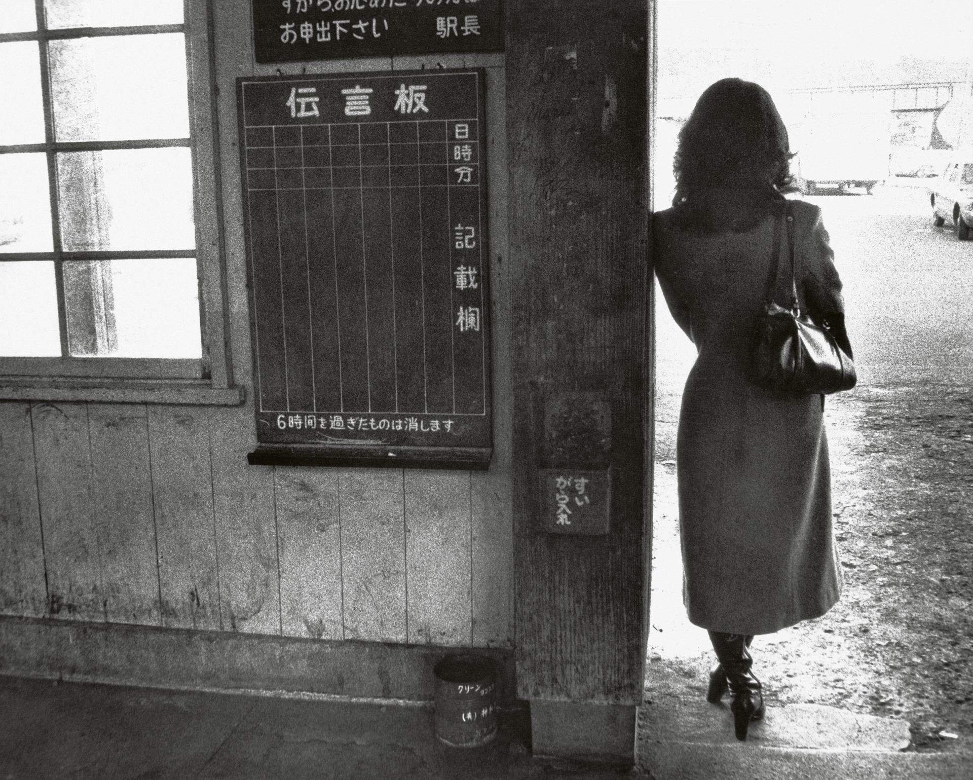 Black and white photograph of a woman standing beside a weathered wooden signboard on an urban street.