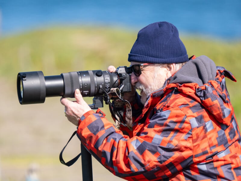 An older man in a red and navy jacket and navy beanie uses a large zoom lens camera outdoors, focusing on a distant subject. The background is blurred, with green and blue tones suggesting nature or a landscape.