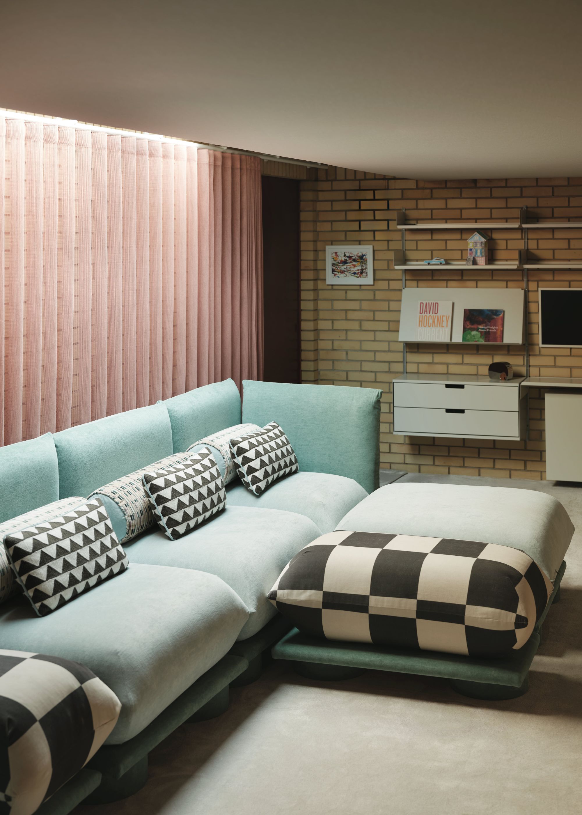 A cinema room in a mid-century home with a turquoise sofa, black and white geometric patterned cushions, a beige carpet, a yellow brick wall, and pink sheer curtains on the other wall.