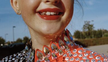 A tight crop of a smiling young dancer's face and throat, her red lips and gap-toothed grin set above a spectacular orange costume collar encrusted with large iridescent crystal gems.