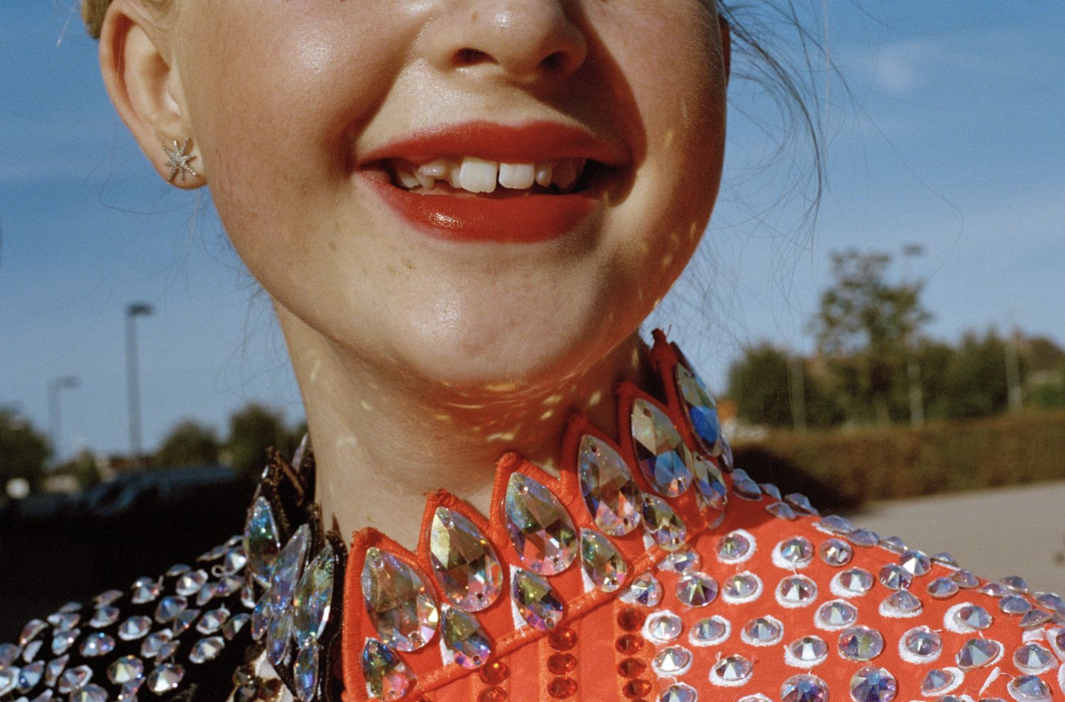 A tight crop of a smiling young dancer's face and throat, her red lips and gap-toothed grin set above a spectacular orange costume collar encrusted with large iridescent crystal gems.
