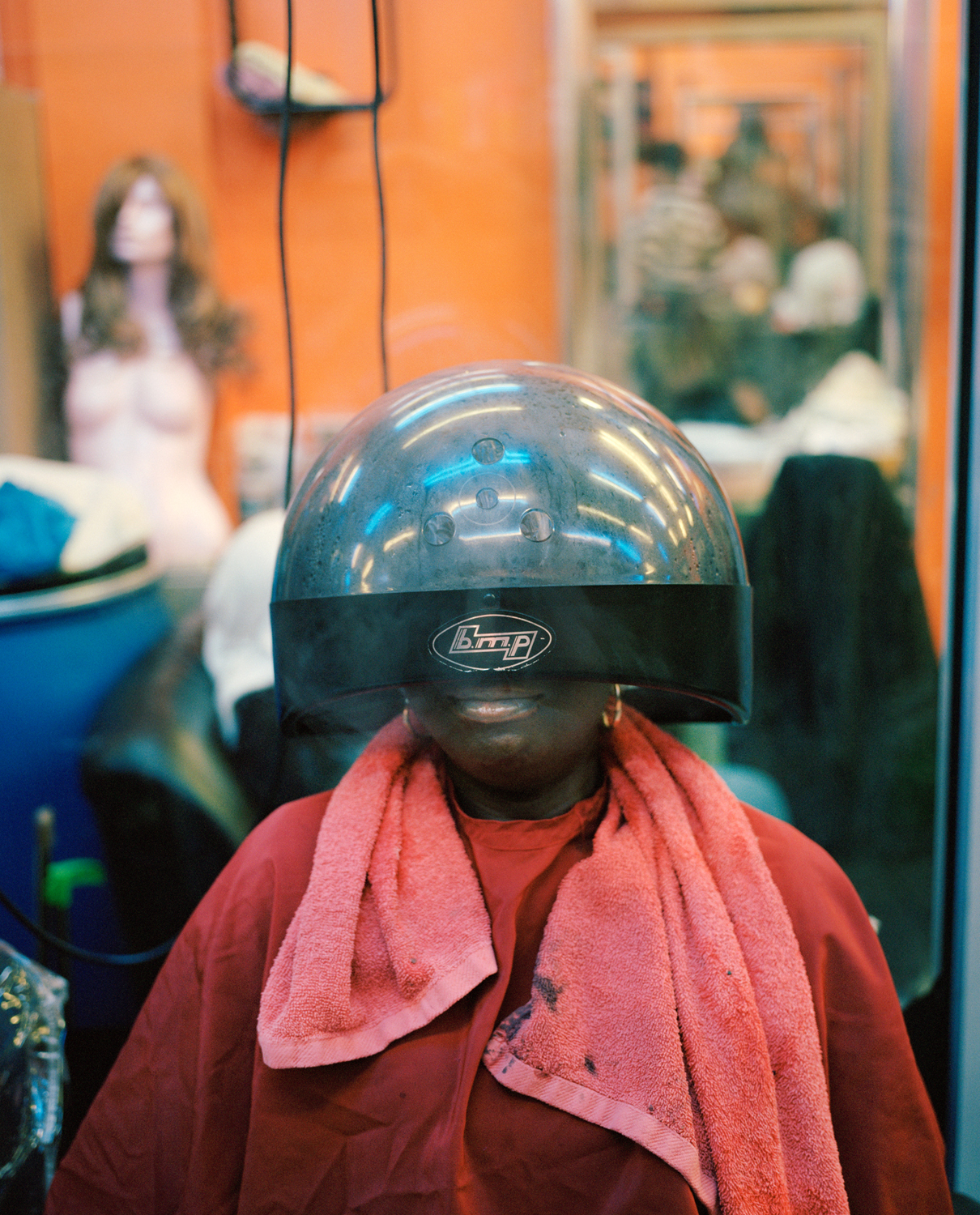 A person sits smiling beneath a large dome hood dryer in a warm amber-toned Peckham salon, a pink towel around their shoulders and a wig mannequin blurred in the background.