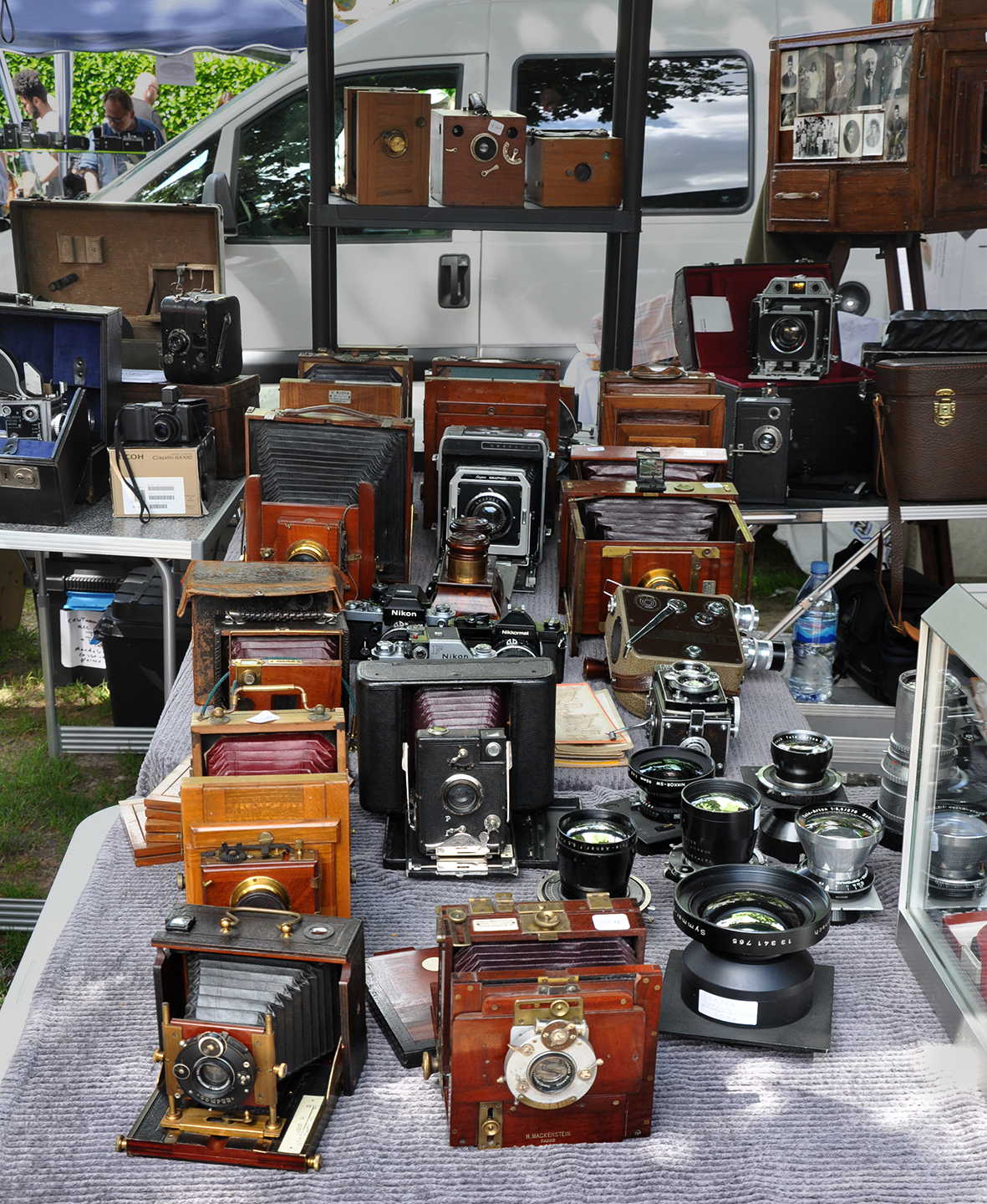 A table crowded with antique cameras spanning more than a century, from brass-fitted wooden plate cameras and bellows folders to mid-century Nikons and a collection of large-format lenses.