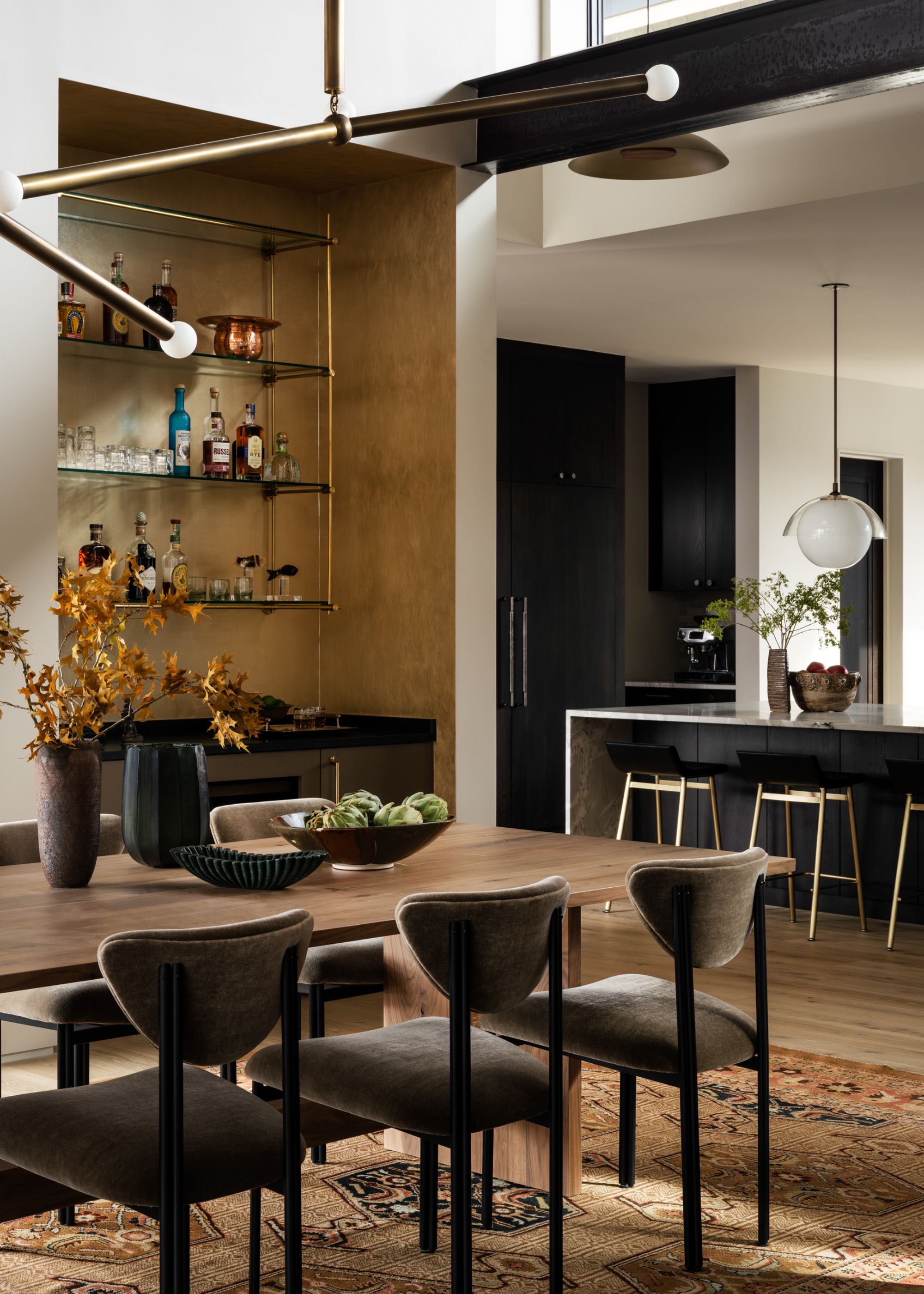 A modern dining space with a home bar in a niche with brass detailing and glass shelving next to the kitchen's island
