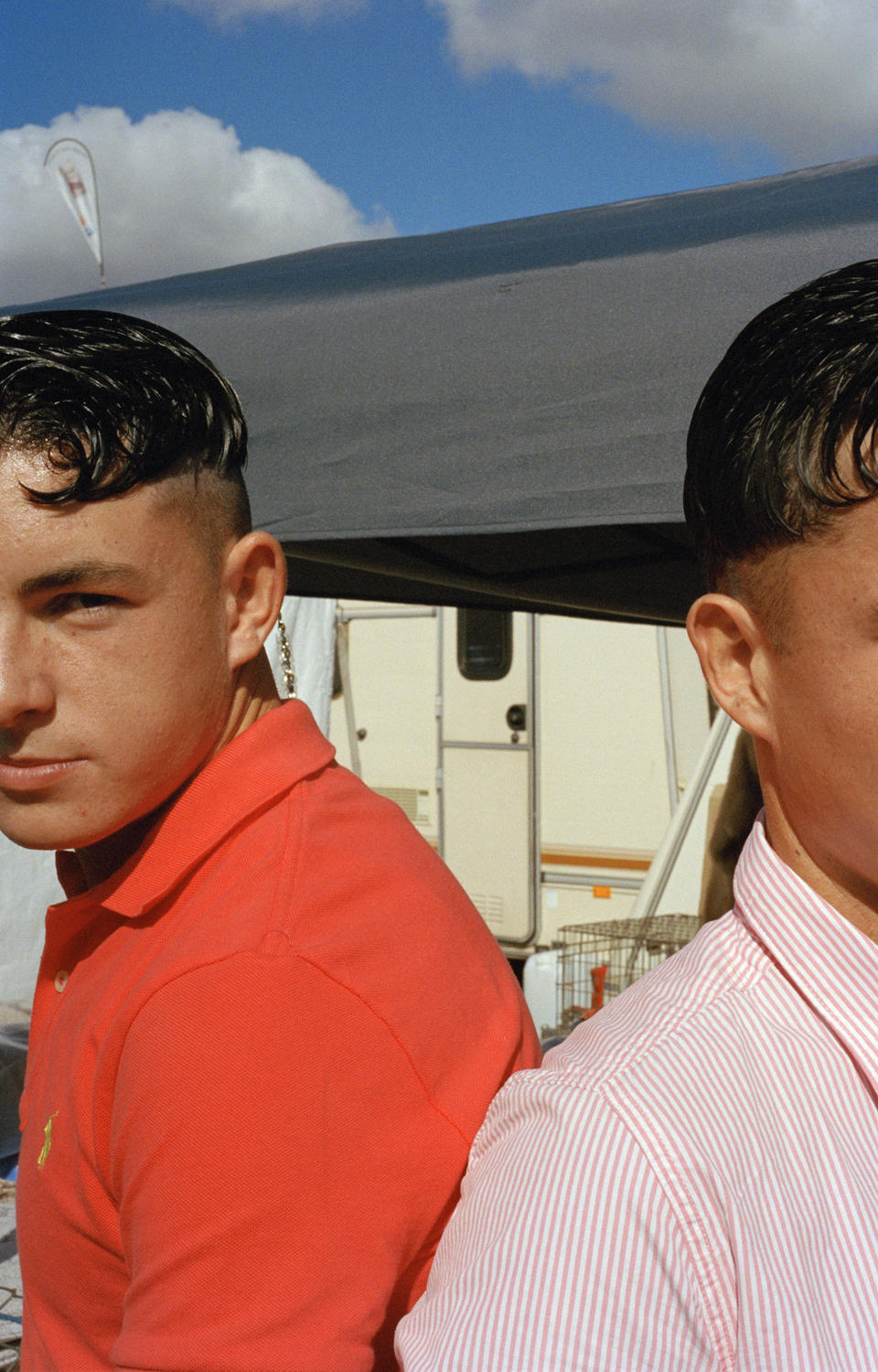 Two young men with identical dark, heavily gelled curls stand shoulder to shoulder at Kenilworth Horse Fair, one in a red polo shirt and one in a pink stripe, a caravan visible behind them.