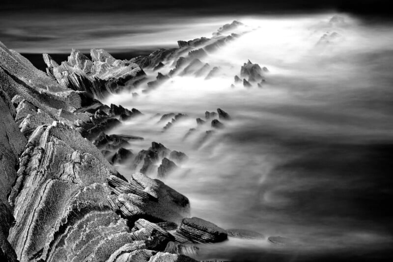 Black-and-white photo of jagged rock formations extending into the sea, with misty water swirling around the rocks, creating a dramatic and ethereal atmosphere.