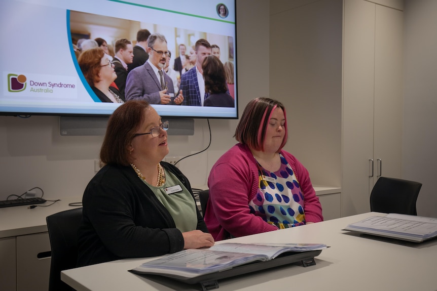 Two women sit at a desk in a training room in front of a screen showing slides for a presentation on health inclusion