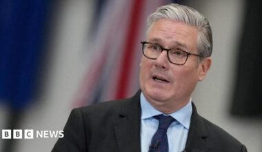 Sir Keir Starmer speaks, wearing spectacles, a dark suit, light blue shirt and a navy blue tie, against a blurred background