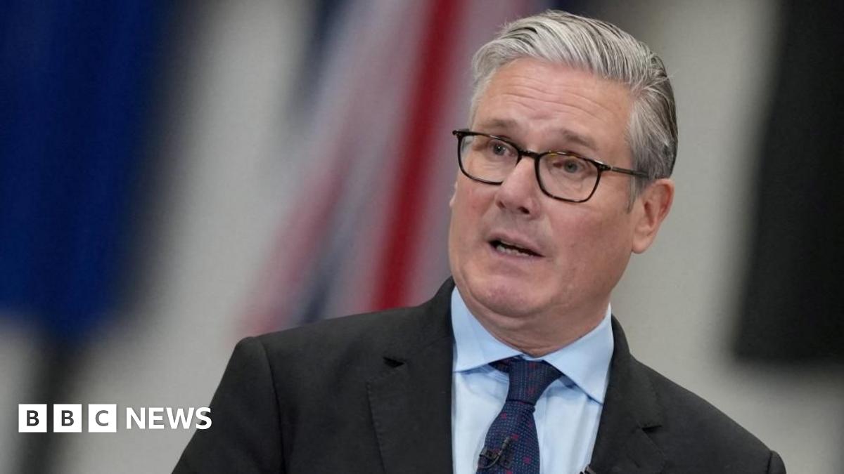 Sir Keir Starmer speaks, wearing spectacles, a dark suit, light blue shirt and a navy blue tie, against a blurred background