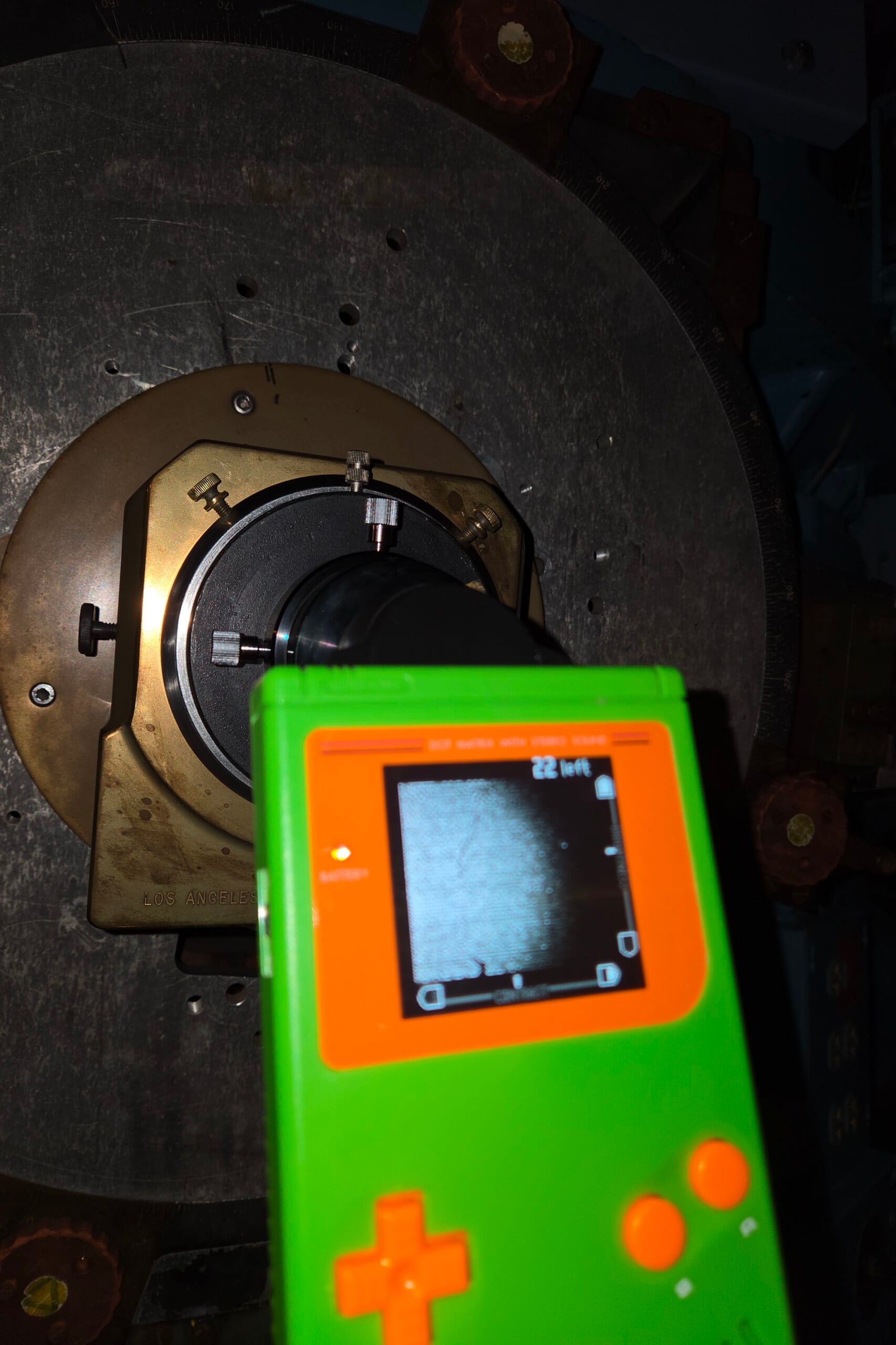 A bright green handheld device with an orange cross button displays a grayscale circular image on its screen, positioned in front of a large, metallic, circular machine component.