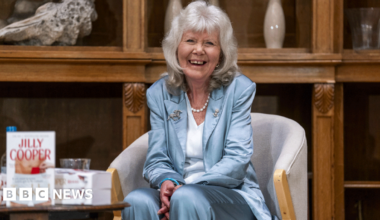 Dame Jilly Cooper is wearing a blue satin suit and stands alongside actors including David Tennant in a group picture. She is smiling at the camera and has her grey hair lose with a fringe.
