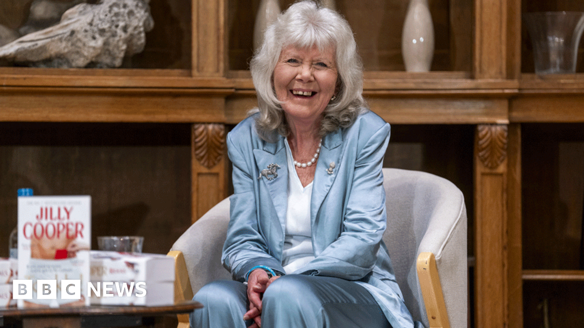Dame Jilly Cooper is wearing a blue satin suit and stands alongside actors including David Tennant in a group picture. She is smiling at the camera and has her grey hair lose with a fringe.
