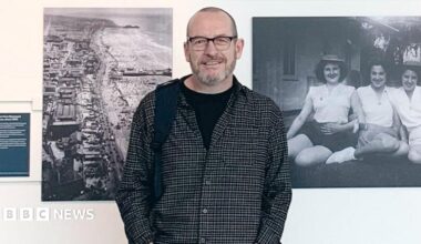 Steven Howells, who is wearing a shirt with a black T-shirt underneath and is smiling at the camera, is standing in front of a white wall with black and white photographs take by his grandad. The picture on the left is an aerial photograph of the coast and the image on the right is on three women sitting on the grass in front of a house.