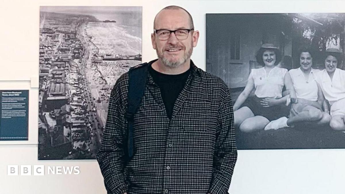 Steven Howells, who is wearing a shirt with a black T-shirt underneath and is smiling at the camera, is standing in front of a white wall with black and white photographs take by his grandad. The picture on the left is an aerial photograph of the coast and the image on the right is on three women sitting on the grass in front of a house.