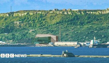 Breakwater with sea either side a large block-shaped building with a chimney in front of steep green cliffs
