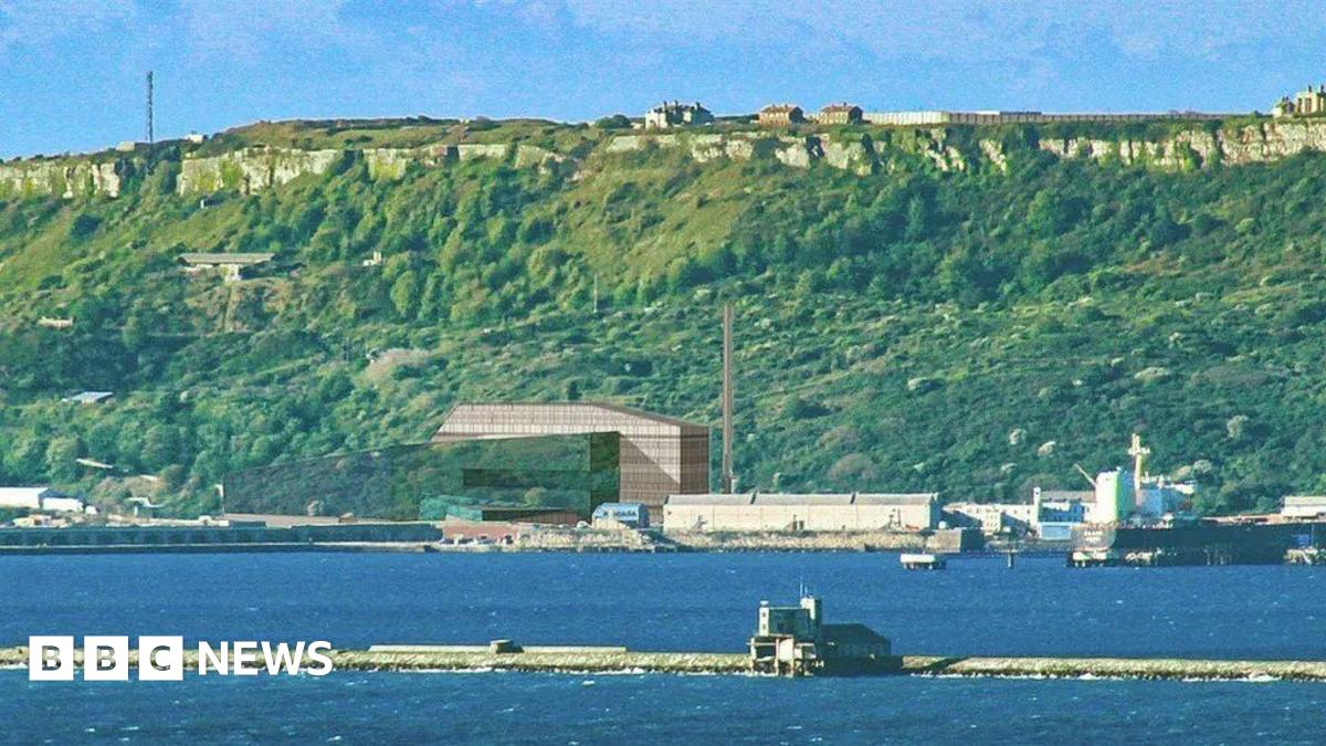 Breakwater with sea either side a large block-shaped building with a chimney in front of steep green cliffs