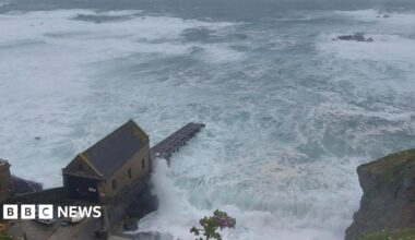 A view of the sea from the top of a cliff. It shows large swells of water and waves crashing against the base of the cliffs. A small building and a slipway are visible.