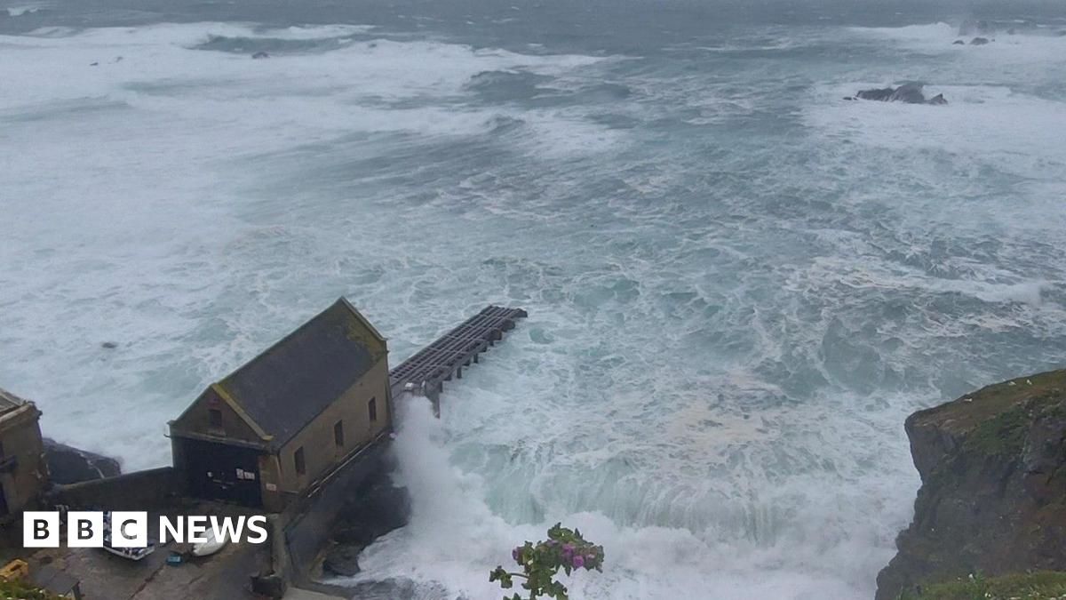 A view of the sea from the top of a cliff. It shows large swells of water and waves crashing against the base of the cliffs. A small building and a slipway are visible.