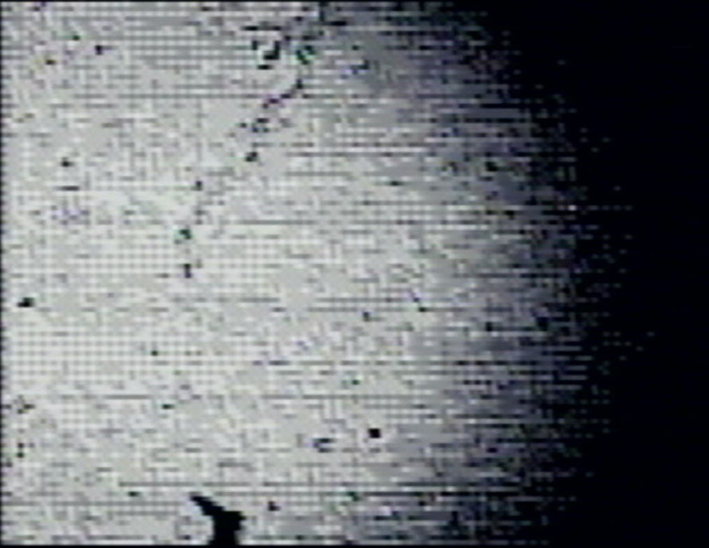 Grainy black-and-white image showing a person in silhouette at the bottom left, appearing to point upward toward a lit, textured wall with visible speckles and a faint vertical crack or line.