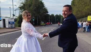 A woman, with blonde hair, tied back, wearing a white wedding dress. She is holding her husband's hand, who is wearing a black wedding suit. They are standing in the middle of a road.