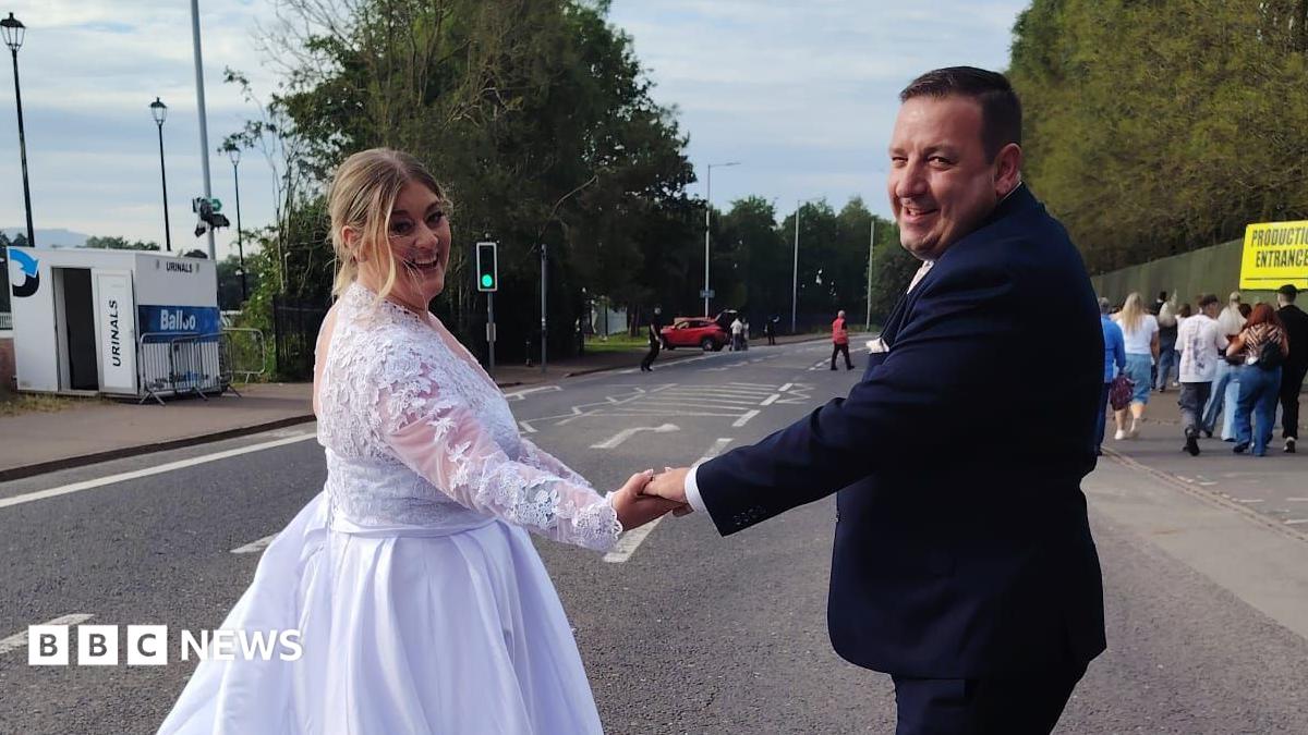 A woman, with blonde hair, tied back, wearing a white wedding dress. She is holding her husband's hand, who is wearing a black wedding suit. They are standing in the middle of a road.