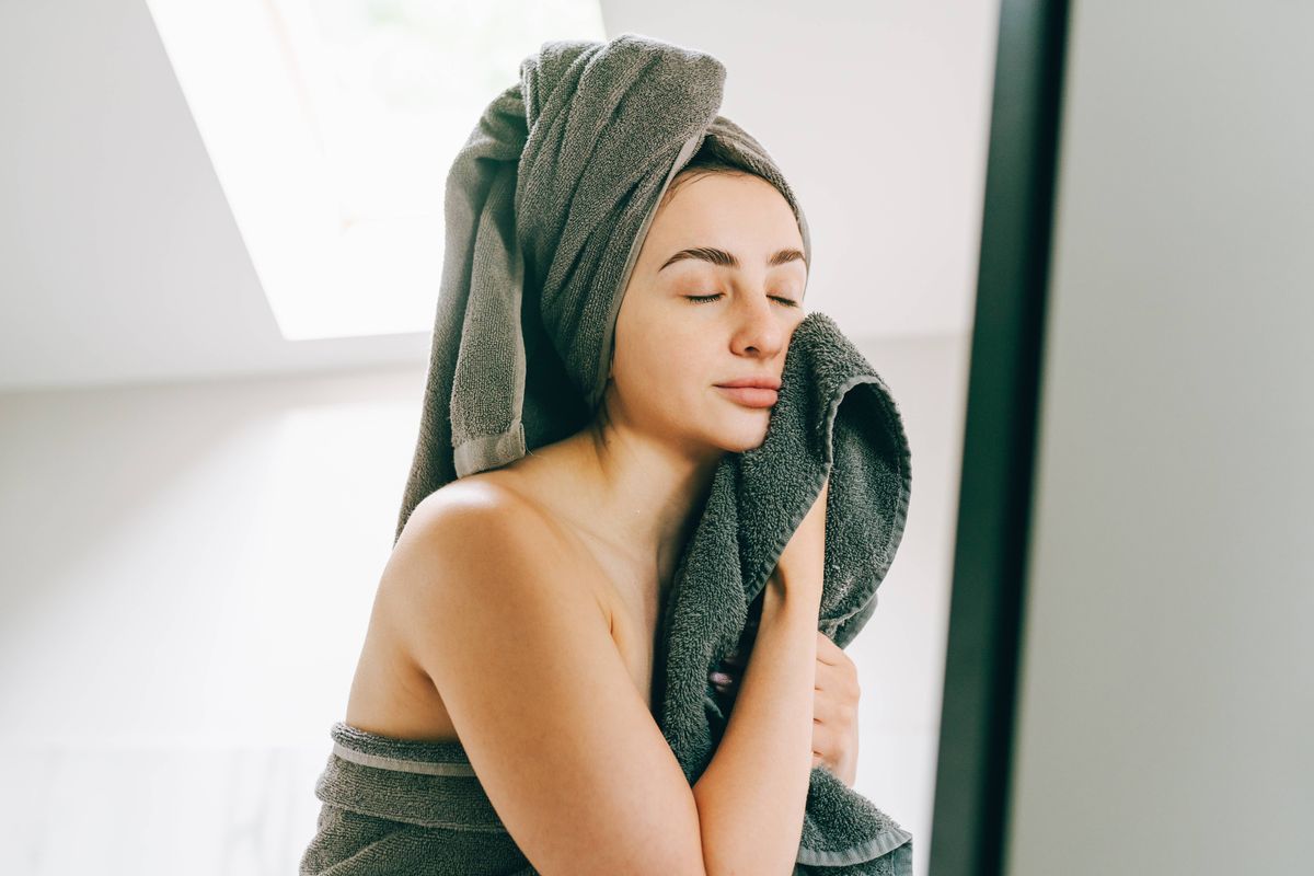 Tranquil woman wrapped in bath towel wiping her face in front of the mirror. Morning skin care and getting ready