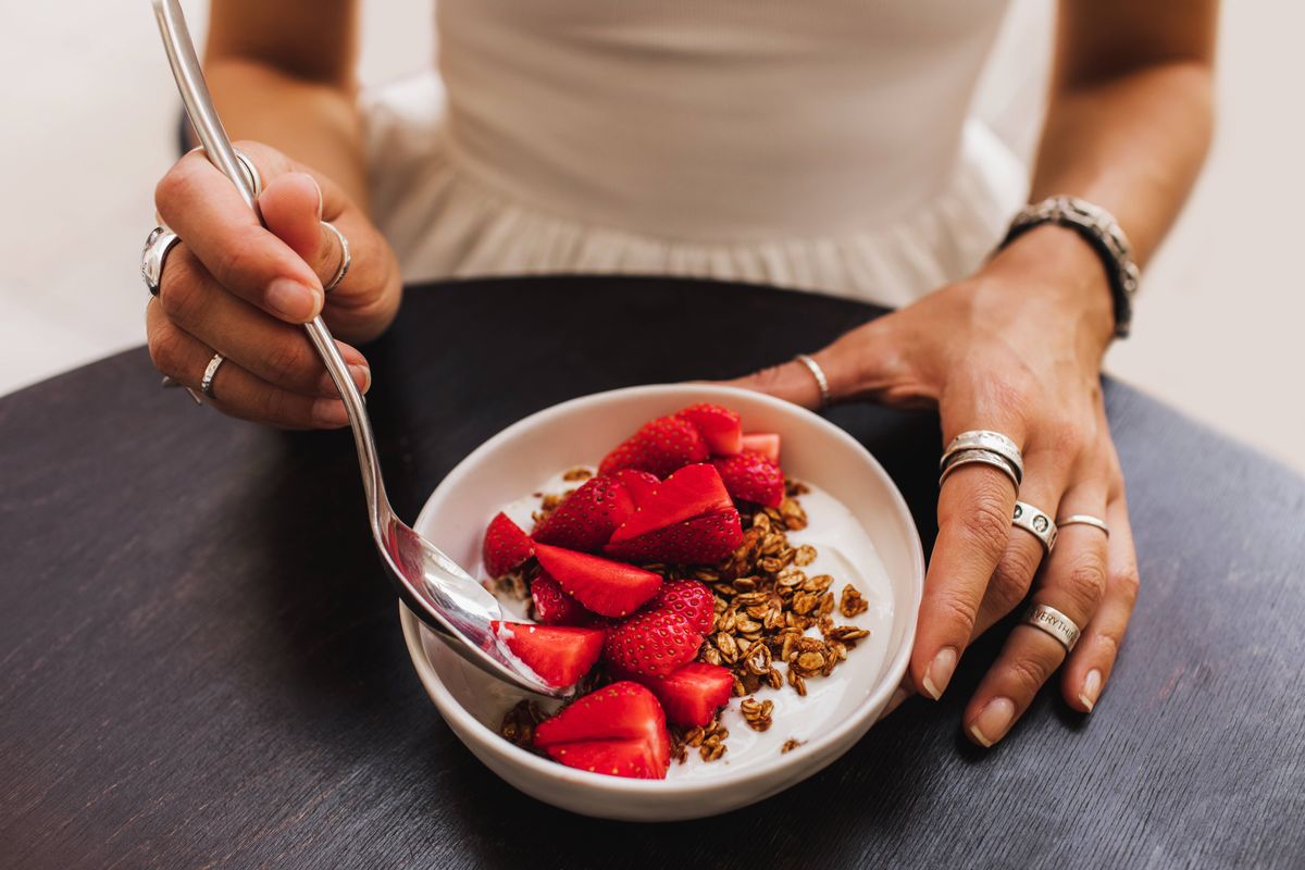 Delicious and nutritious meal served in a white bowl featuring creamy yogurt, crunchy granola, and fresh sliced strawberries, emphasized by human hands holding a spoon on a black tabletop.