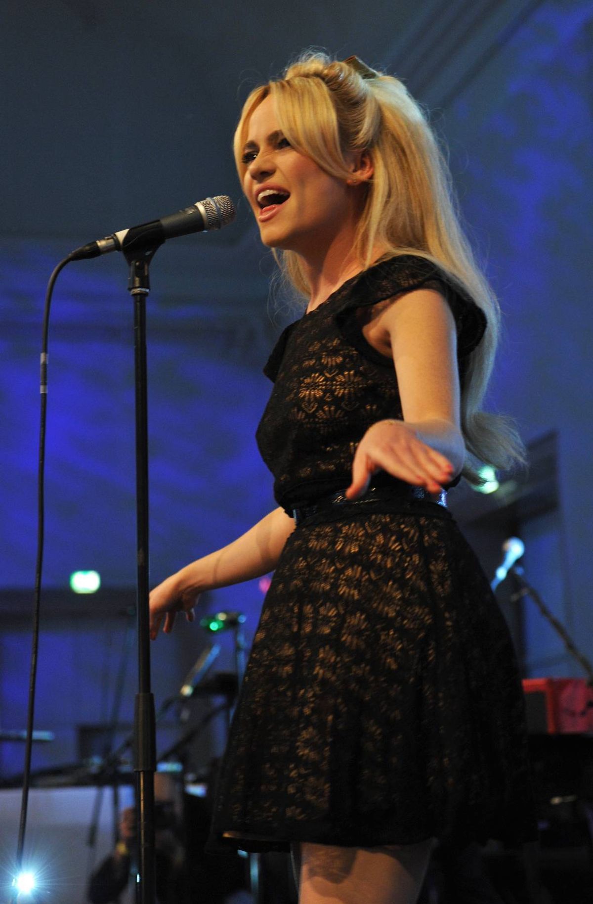 An individual with long blonde hair is performing on stage, singing into a microphone. The setting includes a dark blue backdrop, and the performer is dressed in a black top.