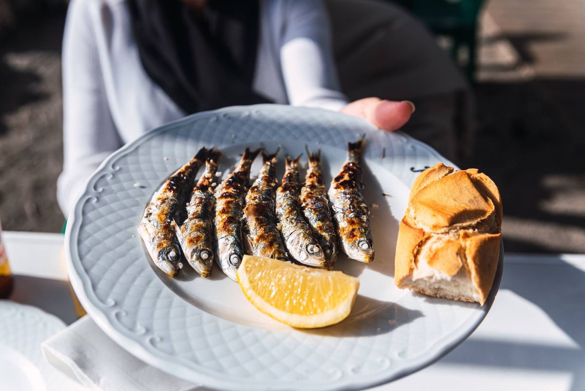 A white plate on a table holds a selection of grilled fish accompanied by a wedge of lemon and a piece of bread.