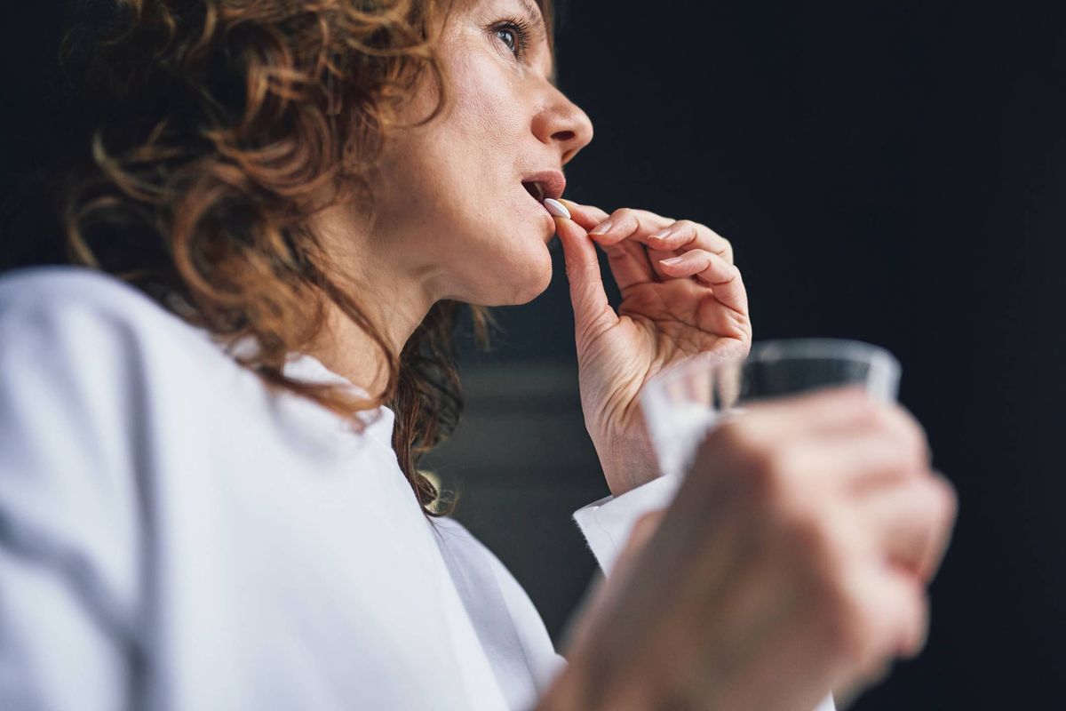An individual dressed in a white long-sleeve shirt is in the act of consuming a pill with the assistance of a clear glass of liquid, captured in a dimly lit setting.
