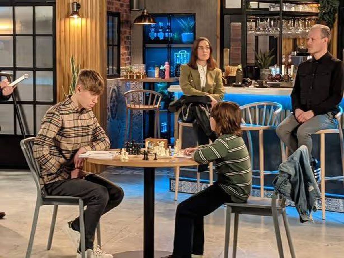 In an indoor setting that resembles a bar or cafe, a group of individuals are engaged in various activities. Two individuals are seated at a table, deeply engrossed in a game of chess, with a few pieces strategically placed on the board. Behind them, another individual is standing, holding a microphone, possibly conducting an interview or engaging in conversation. A fourth person is seated on a stool, attentively observing the scene. The ambiance is enhanced by the presence of a bar counter with several glasses and other items. The overall setting appears casual yet focused on the activities at hand.